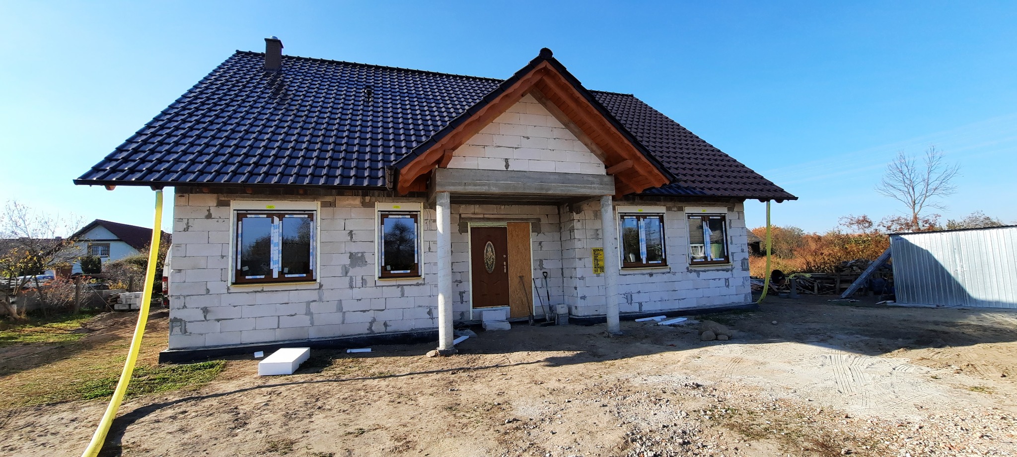 Surowy stan budynku jednorodzinnego z ciemnobrązową dachówką, brązowymi ramami okiennymi i częściowo zamontowanymi oknami, widoczne bloczki betonowe i przygotowane wejście.