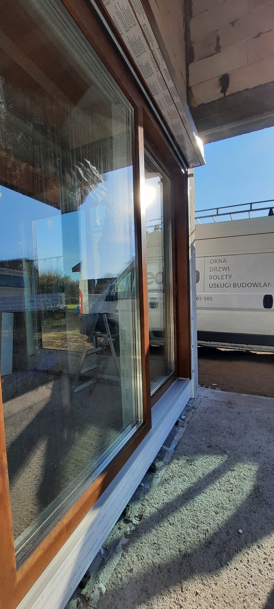 Montaż brązowych ram okiennych z widocznym parapetem zewnętrznym, na tle częściowo widocznego samochodu dostawczego z logo firmy budowlanej.