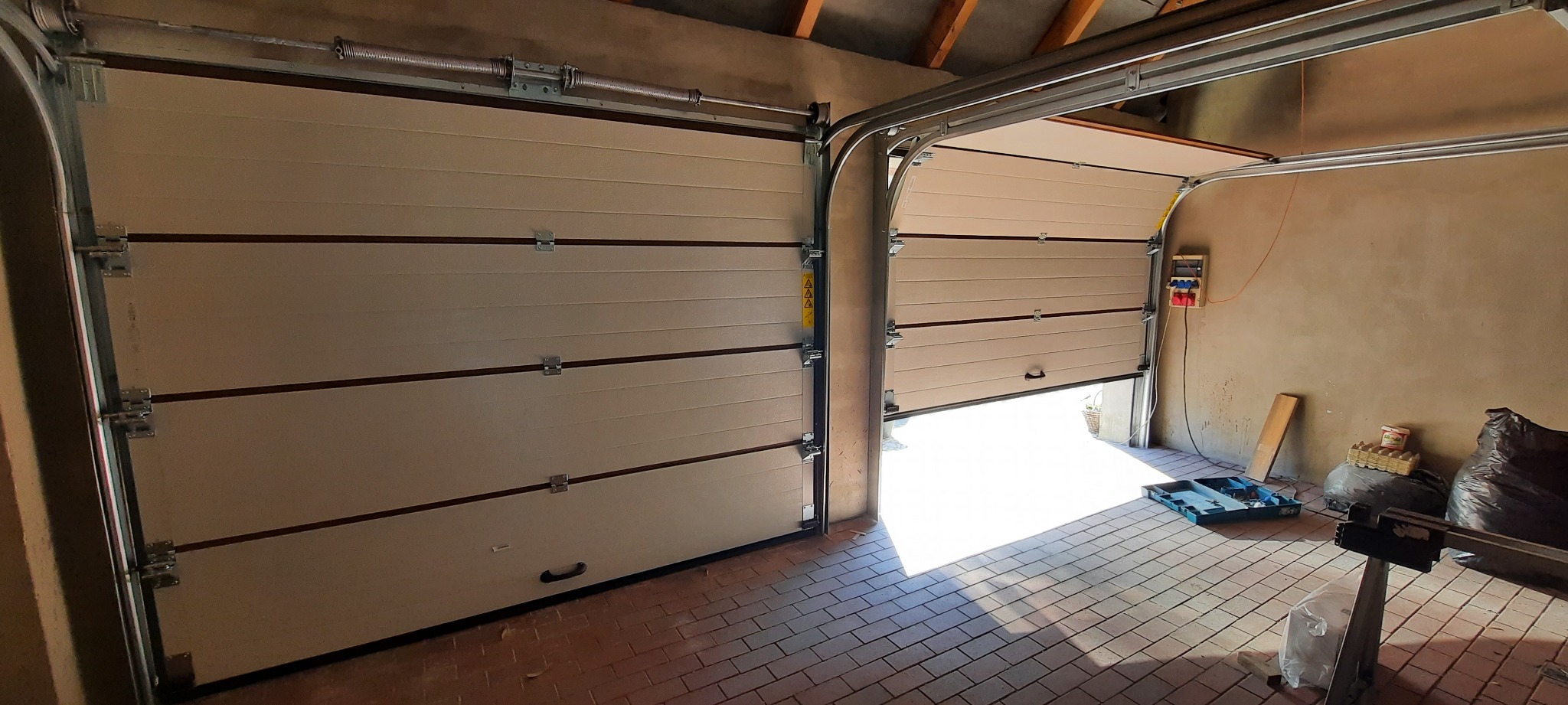 Otwarta segmentowa brama garażowa w kolorze beżowym, widoczne prowadnice i mechanizm sprężynowy, ceglana podłoga garażu z narzędziami i akcesoriami w tle.