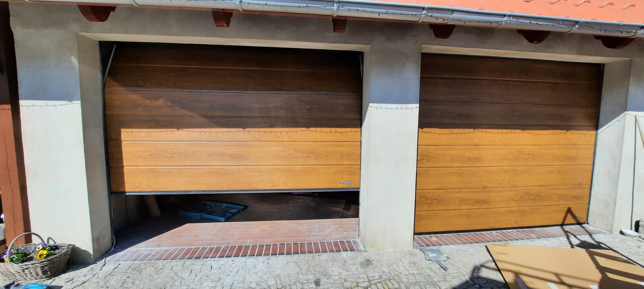 Dwa segmentowe brązowe drzwi garażowe z imitacją drewna, jedno częściowo otwarte, widoczny mur z cegieł i narzędzia wewnątrz garażu, na zewnątrz kostka brukowa i koszyk z kwiatami.