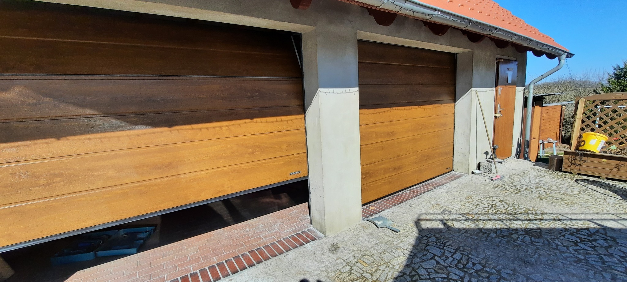 Dwa garaże z segmentowymi bramami w kolorze drewna, jedna brama lekko uchylona, widoczna kostka brukowa i fragment ogrodu w tle.