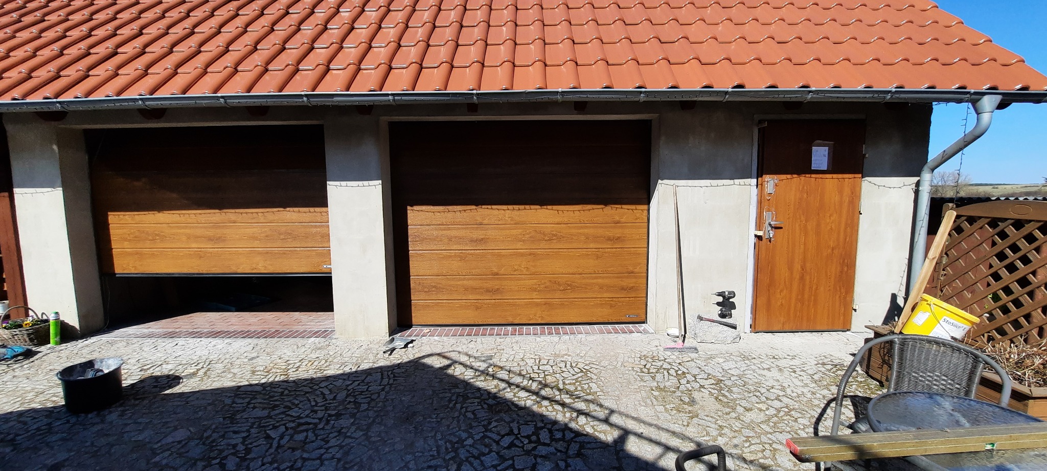 Garaż z dwoma drewnopodobnymi bramami segmentowymi, z których jedna jest częściowo otwarta, oraz brązowymi drzwiami wejściowymi obok, widoczne narzędzia i wiadro z farbą, w tle dach z czerwonej...