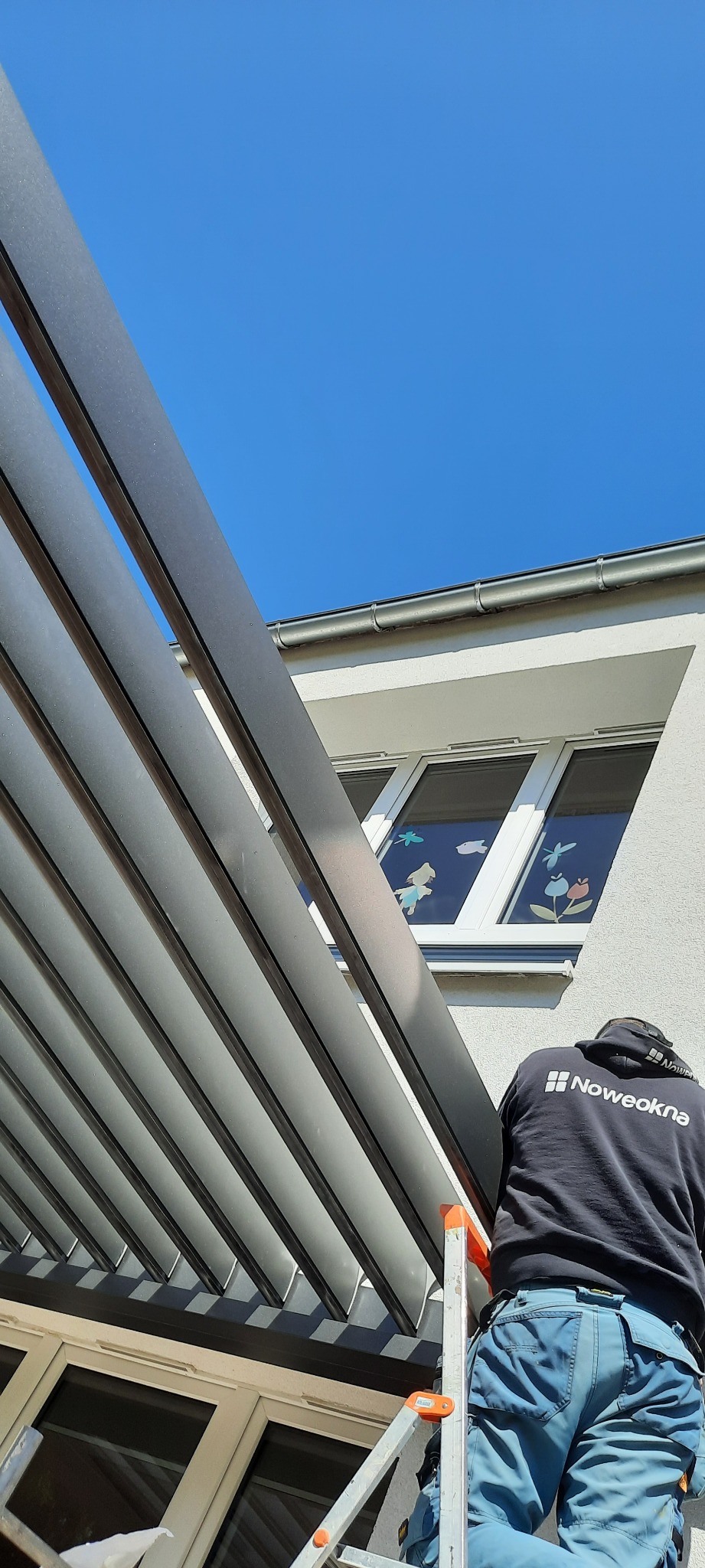 Montażysta na drabinie instaluje aluminiową konstrukcję z regulowanymi lamelami, widoczne okno z naklejkami dziecięcymi, budynek w tle, słoneczny dzień.