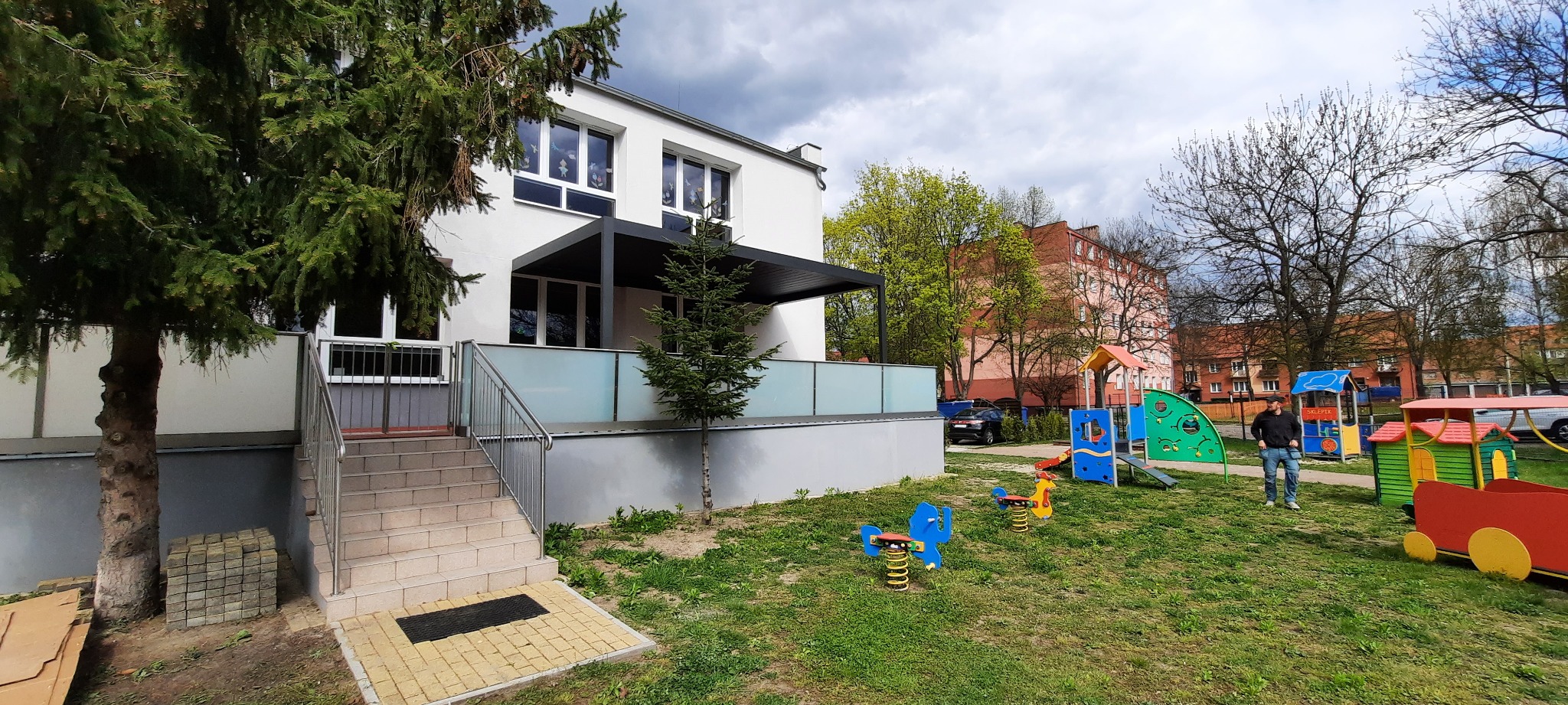 Biały budynek z oknami i szarą pergolą, widok z zewnątrz na plac zabaw z kolorowymi zabawkami i drzewami w tle.