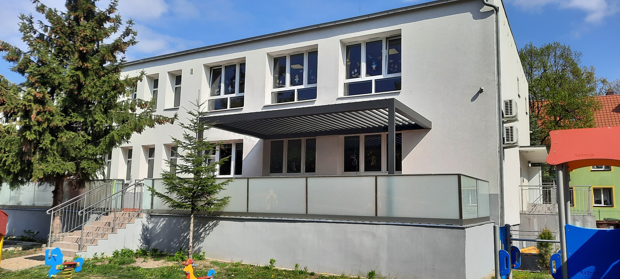 Dwukondygnacyjny budynek z jasną elewacją, oknami z dekoracjami, zadaszeniem lamelowym nad balkonem z mlecznym szkłem, widok z placu zabaw.