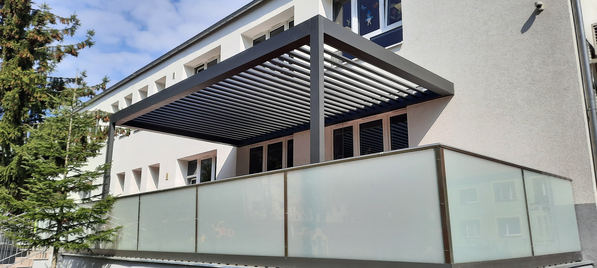 Nowoczesna pergola z poziomymi lamelami w kolorze antracytowym, zamontowana nad balkonem z matowym szkłem, widok z zewnątrz na tle jasnej elewacji budynku i zieleni drzew