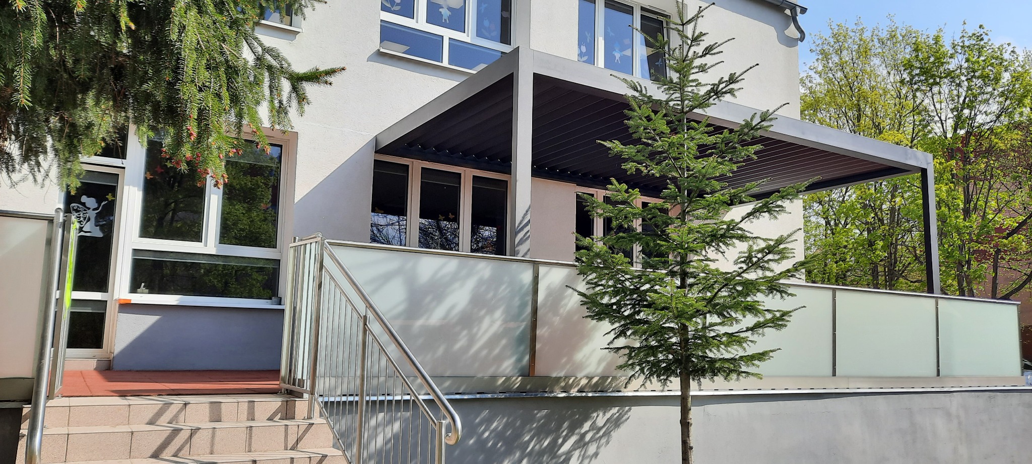 Nowoczesna pergola tarasowa z żaluzjami regulowanymi, widok z zewnątrz, z ozdobnym drzewkiem na pierwszym planie i oknami z naklejkami w tle.