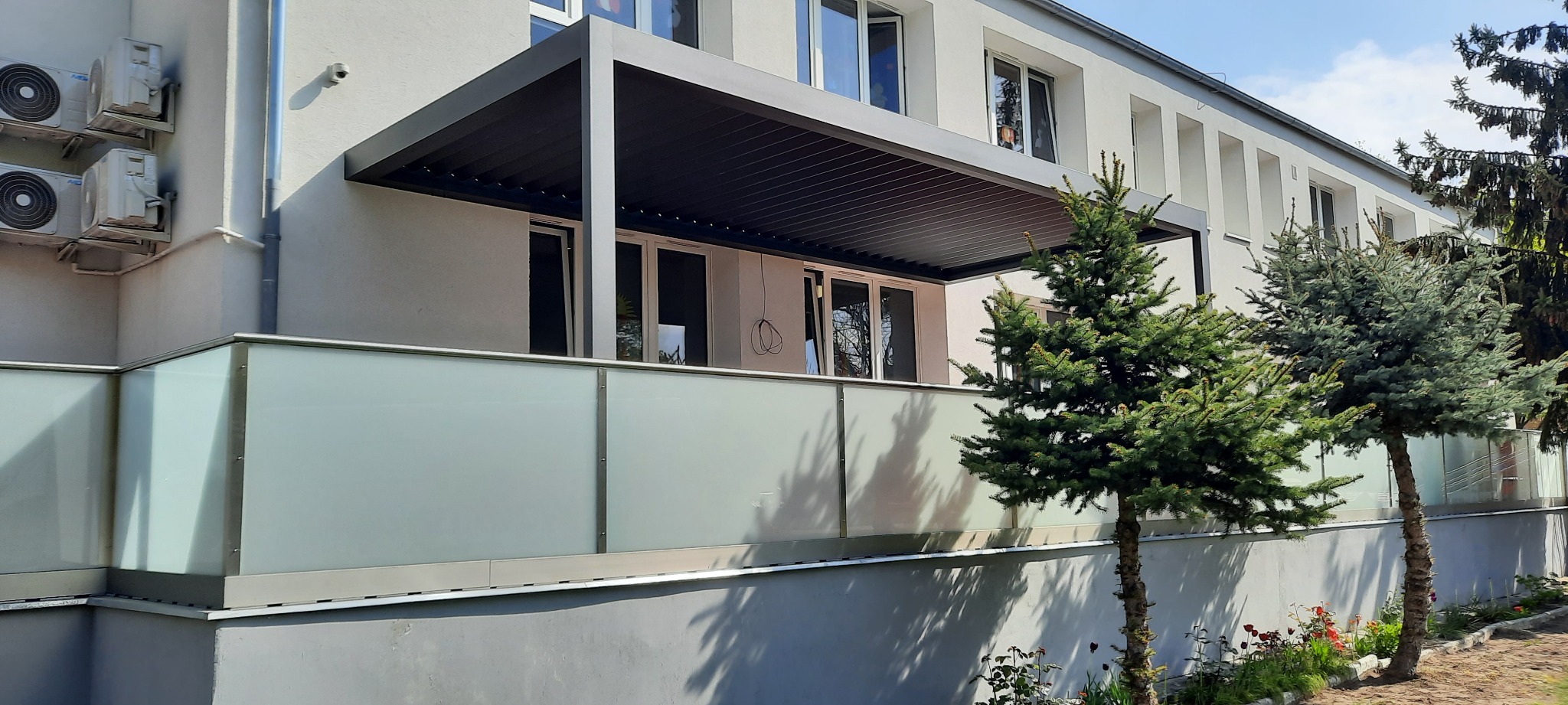 Nowoczesny balkon z szklaną balustradą i zadaszeniem w kolorze antracytowym, widok z zewnątrz na tle budynku z klimatyzatorami i dwoma młodymi drzewkami iglastymi.