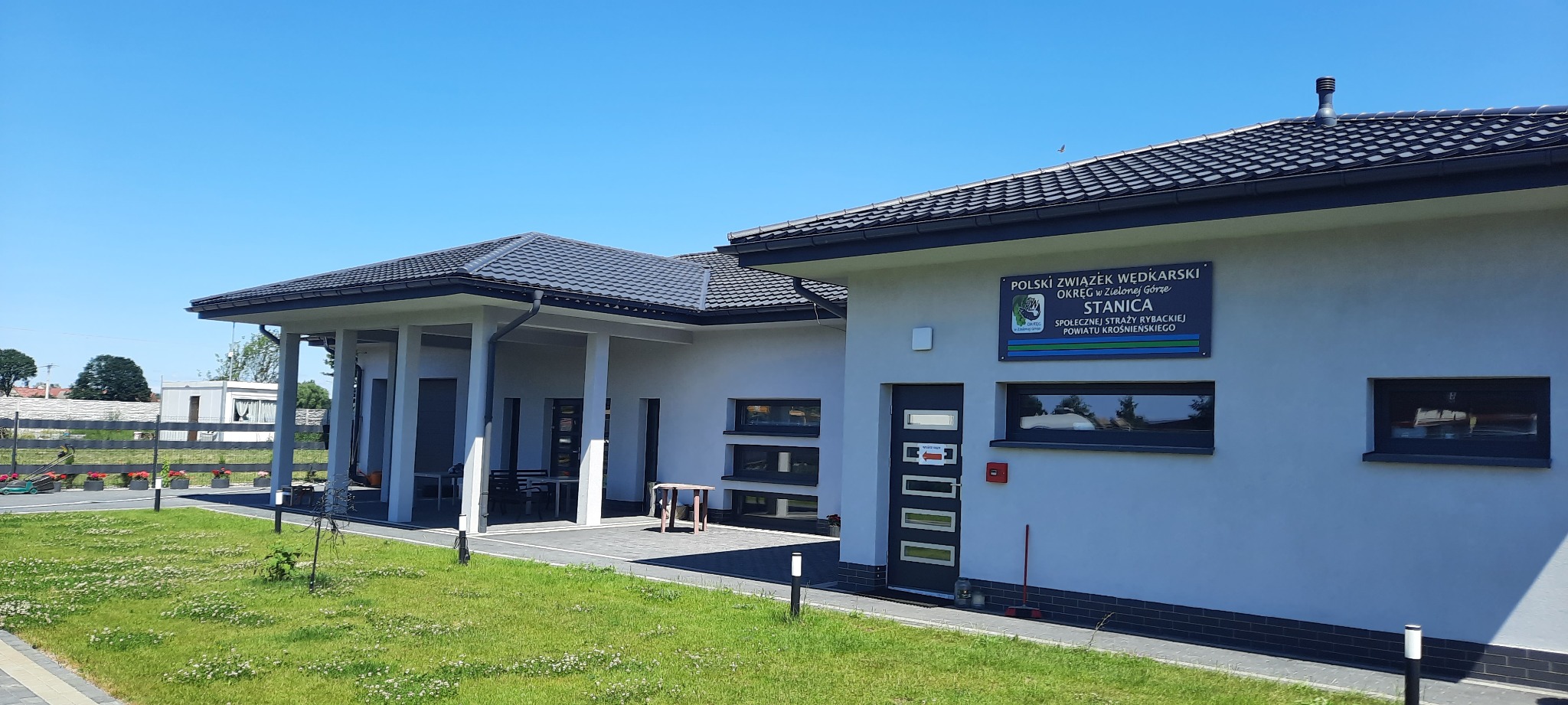 Budynek stanicy wędkarskiej z oknami PCV, ciemną dachówką i starannie utrzymanym trawnikiem w słoneczny dzień.