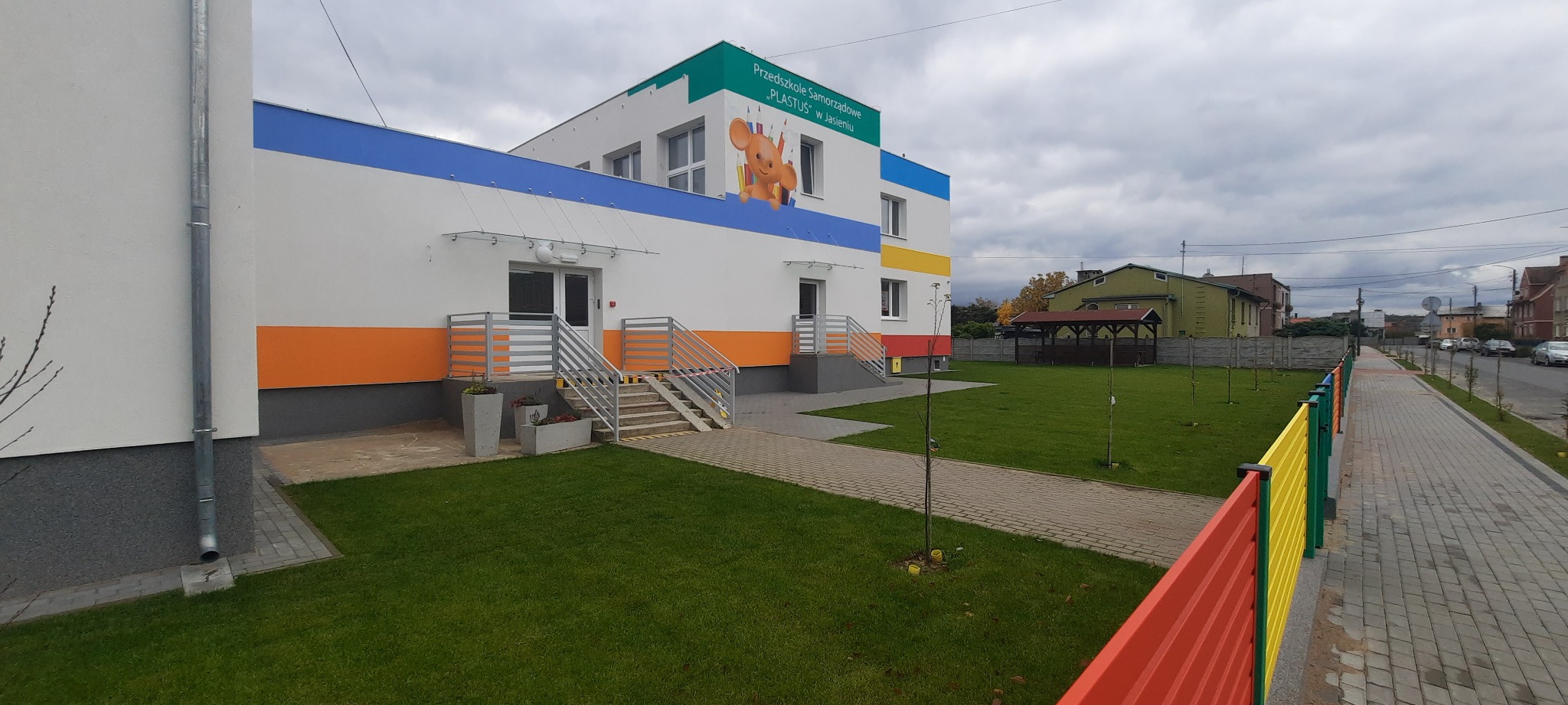 Nowo wybudowany budynek przedszkola z białymi ścianami, kolorowymi pasami (niebieski, pomarańczowy, żółty) i szarymi schodami wejściowymi, otoczony zielonym trawnikiem i chodnikiem z kostki brukowej.