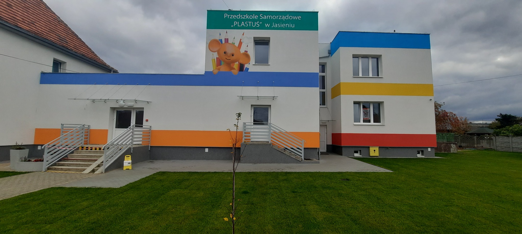 Nowy budynek przedszkola z kolorowymi pasami (niebieski, żółty, czerwony, pomarańczowy) i logo Plastusia na elewacji