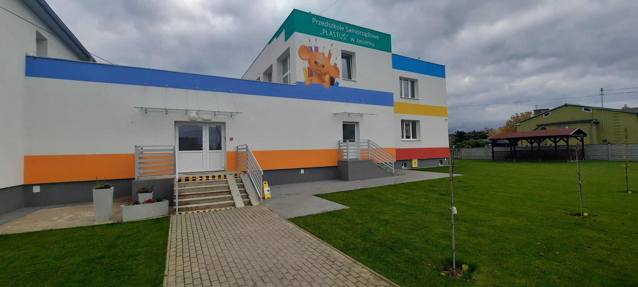 Fasada budynku przedszkola z białymi drzwiami wejściowymi, rampą dla wózków, kolorowymi pasami na elewacji (pomarańczowy, niebieski, żółty, czerwony) i logo z wizerunkiem myszki na tle kredek.