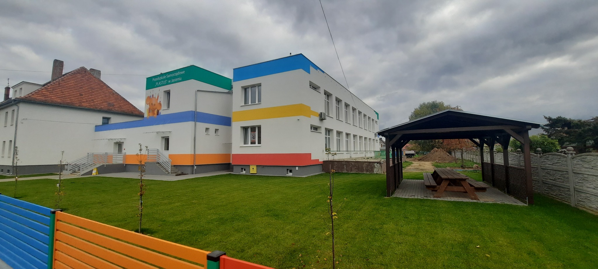 Nowo wybudowany, kolorowy budynek przedszkola z oknami, otoczony trawnikiem i drewnianą altaną z ławkami.