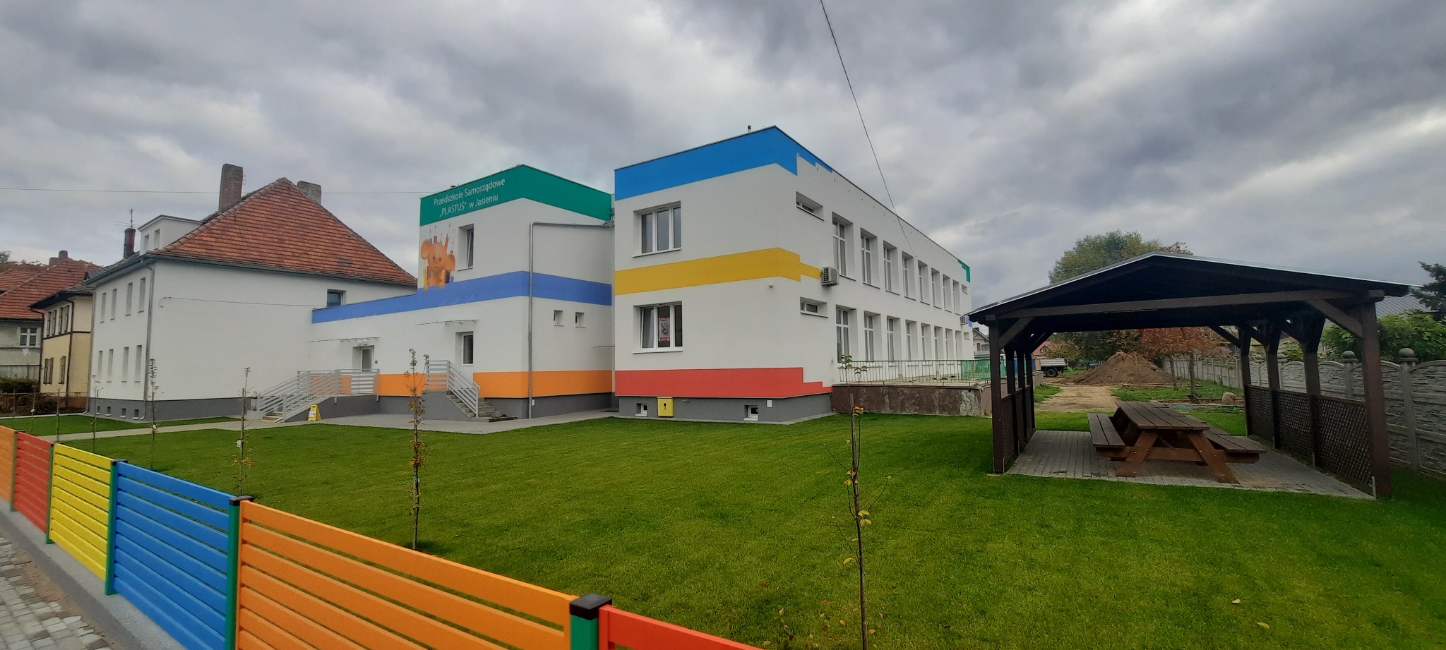 Nowoczesny budynek przedszkola z kolorowymi akcentami, widoczne okna, ogrodzenie z desek w barwach tęczy i drewniana altana z ławkami na trawniku.