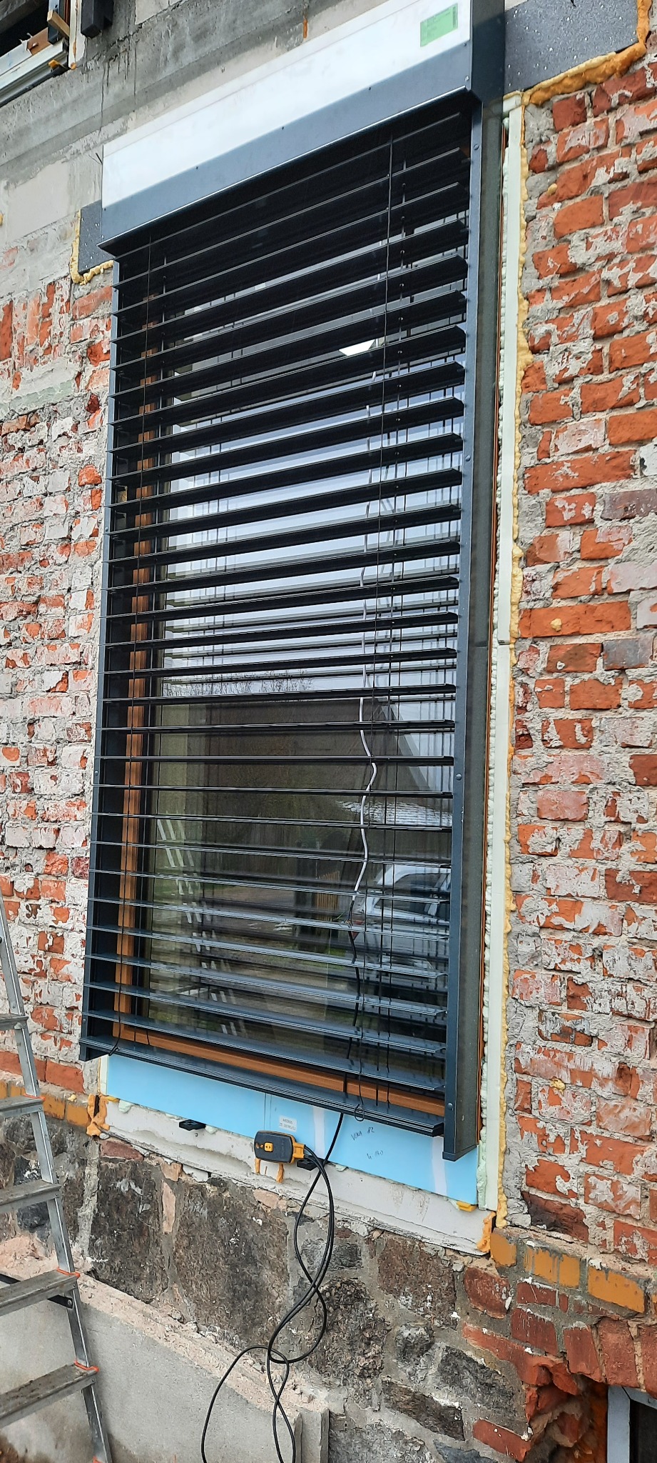Czarne żaluzje fasadowe zamontowane w oknie budynku z cegły, widoczne pianka montażowa i narzędzia instalacyjne.