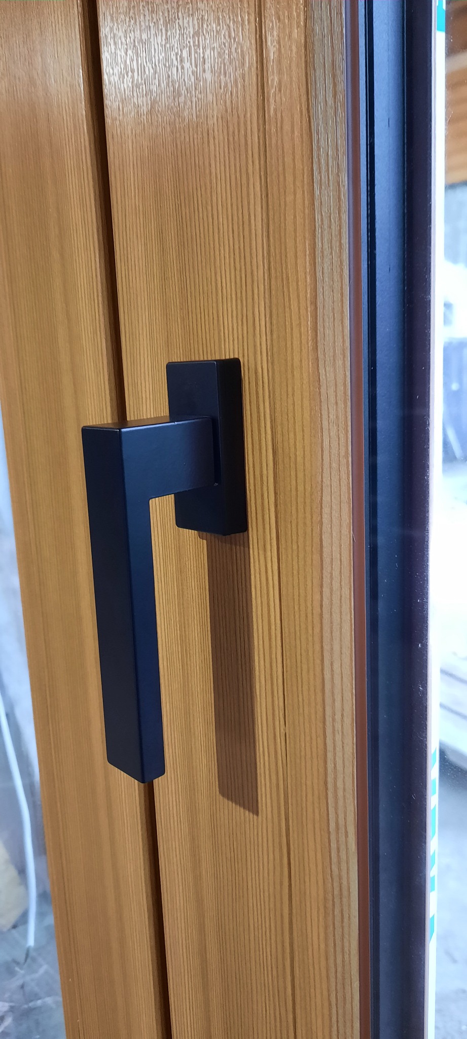 Zamknięte okno z drewna w jasnym odcieniu z czarną, minimalistyczną klamką, widoczna szyba i rama