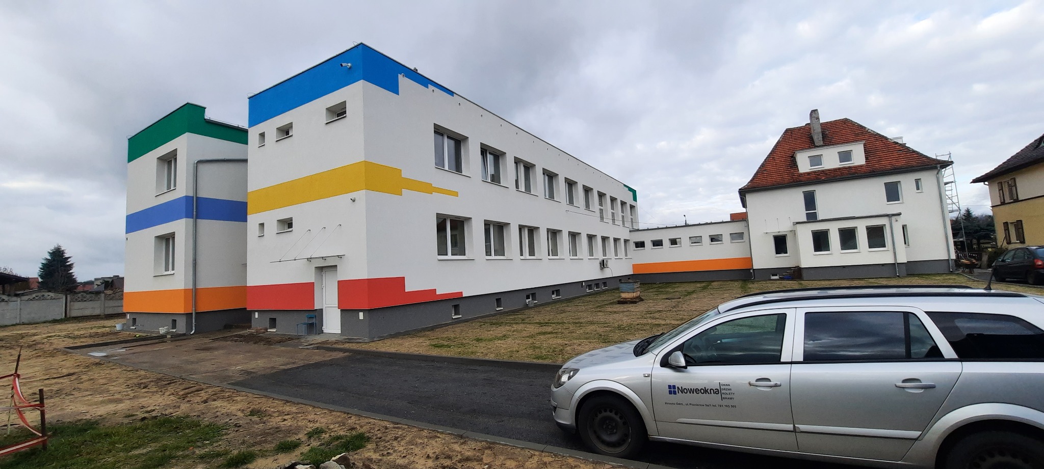 Odnowiony budynek szkoły z kolorowymi pasami na elewacji, widoczne białe okna, szary samochód z logo firmy w pobliżu.