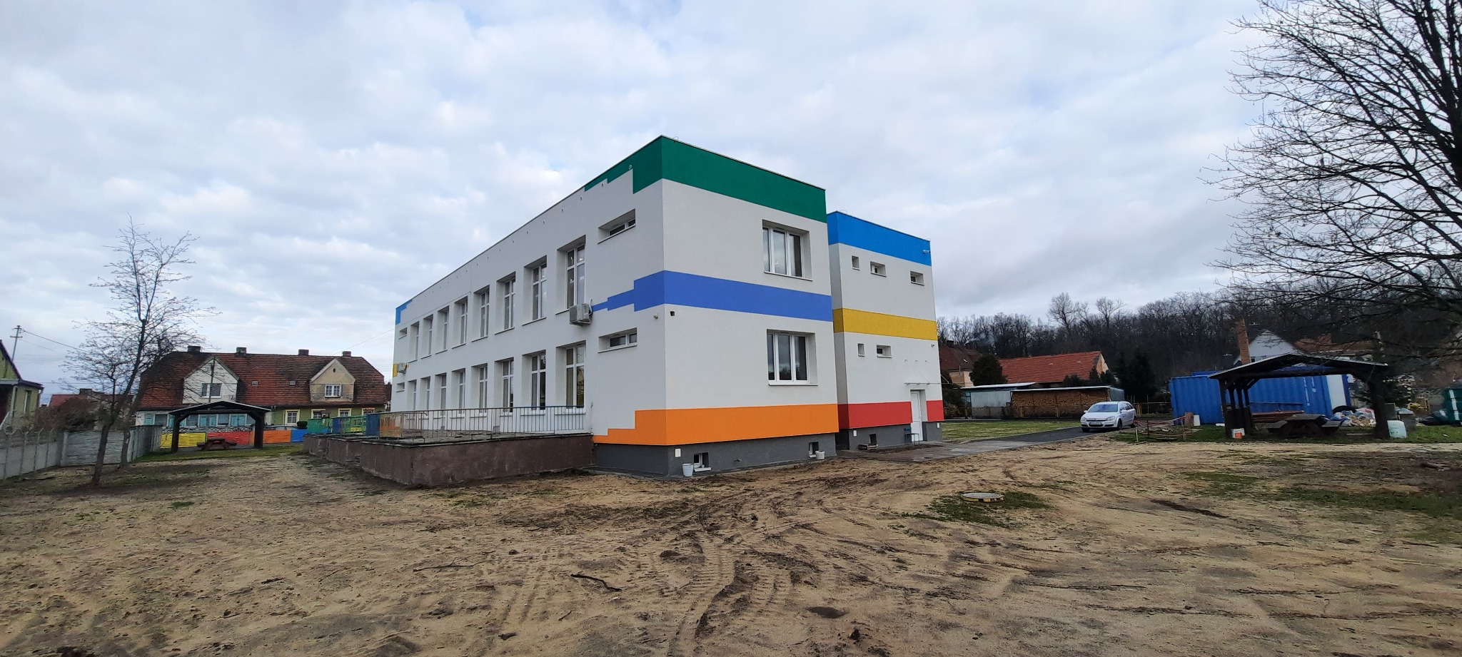 Nowo pomalowany, dwukondygnacyjny budynek z białymi ścianami i kolorowymi pasami (zielony, niebieski, żółty, pomarańczowy, czerwony) pod oknami, widok z zewnątrz na tle pochmurnego nieba i innych...