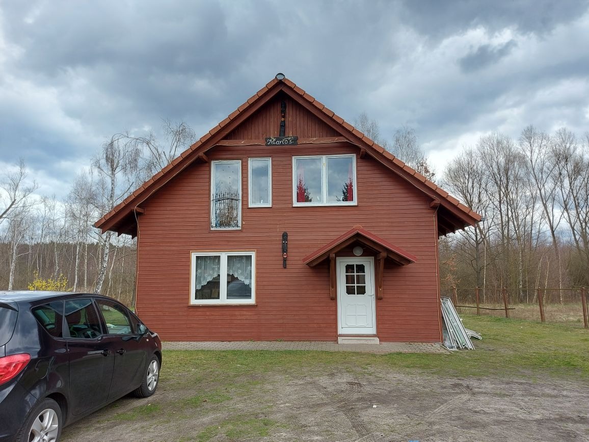 Czerwony dom z białymi oknami i drzwiami wejściowymi, widoczne drzewa w tle i fragment samochodu po lewej stronie.