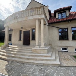 Zakład kamieniarski D&BD - Eleganckie schody kamienne prowadzą do wejścia domu z kolumnami i balkonem. Kamienna kostka brukowa na podjeździe. Całość w jasnych, stonowanych barwach.