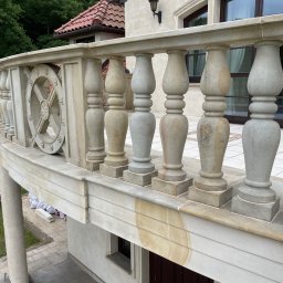 Zakład kamieniarski D&BD - Kamienna balustrada balkonu z dekoracyjnymi elementami, widok z góry na trawnik i pracownika w trakcie montażu. Widoczne detale wykończenia i faktura kamienia.