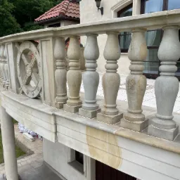 Kamienna balustrada balkonu z dekoracyjnymi elementami, widok z góry na trawnik i pracownika w trakcie montażu. Widoczne detale wykończenia i faktura kamienia.