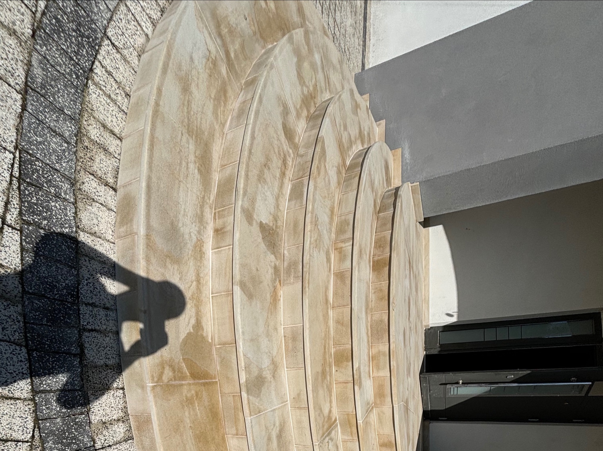 Wykończone schody zewnętrzne z płytek w odcieniach beżu, z widocznym cieniem osoby robiącej zdjęcie. Elementy architektoniczne i fragment okna w tle.