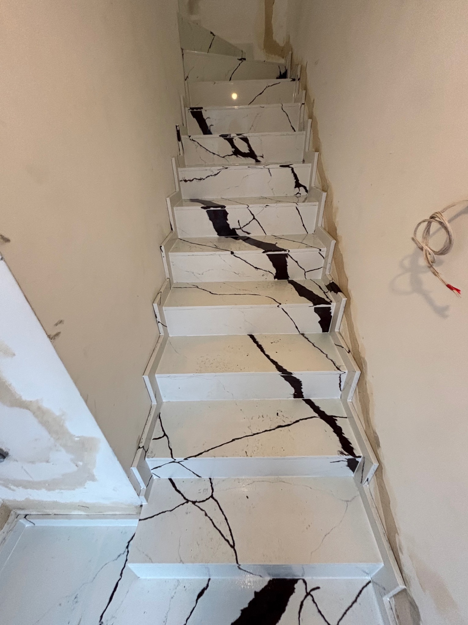 Wykończone schody z białego kamienia z czarnymi żyłami, widok z góry. Ściany w kolorze beżowym, widoczne niedociągnięcia wykończeniowe i wystające przewody.