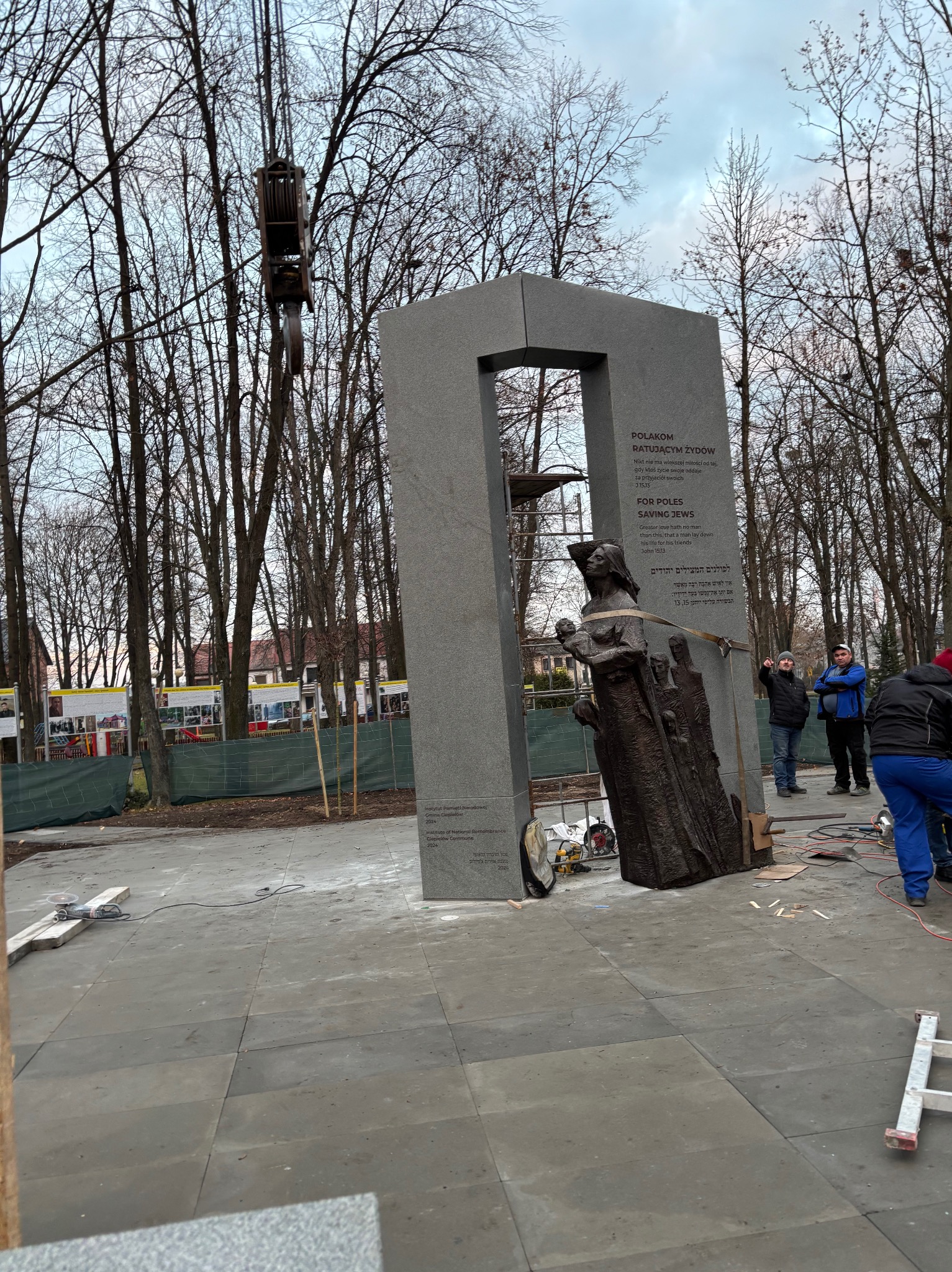 Montaż pomnika w Krakowie: dźwig podnosi rzeźbę z brązu na tle kamiennej konstrukcji z inskrypcjami. Widoczni robotnicy i narzędzia.