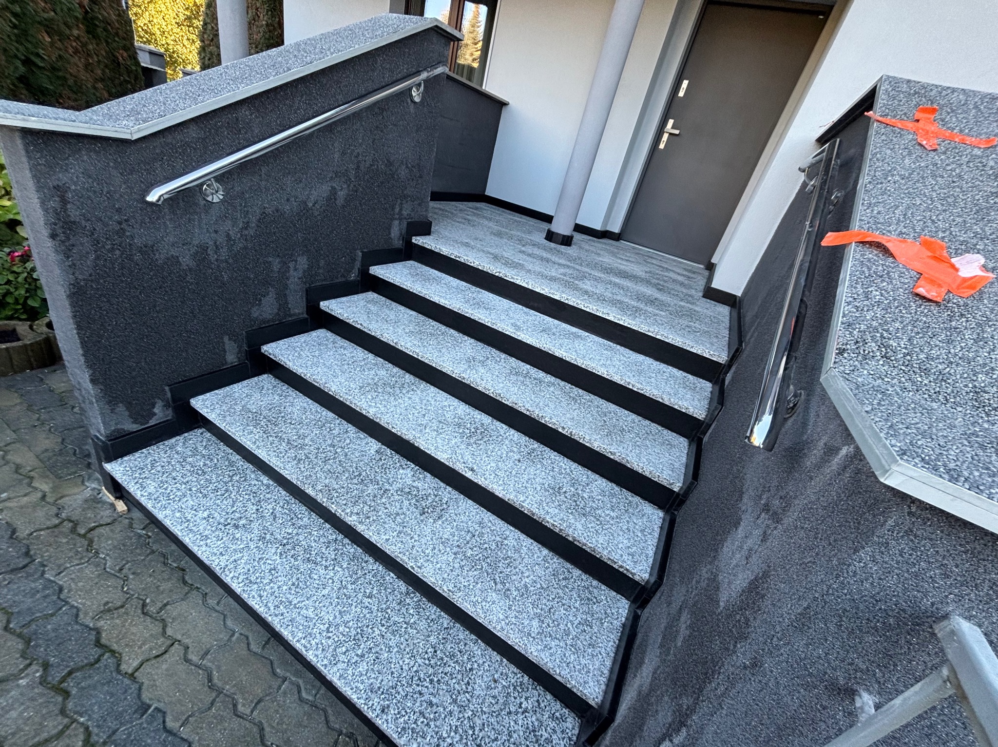 Zewnętrzne schody z szarego kamienia z czarnymi obramowaniami, prowadzące do drzwi wejściowych. Metalowa poręcz po lewej stronie. Widoczne mocowania poręczy do ściany.