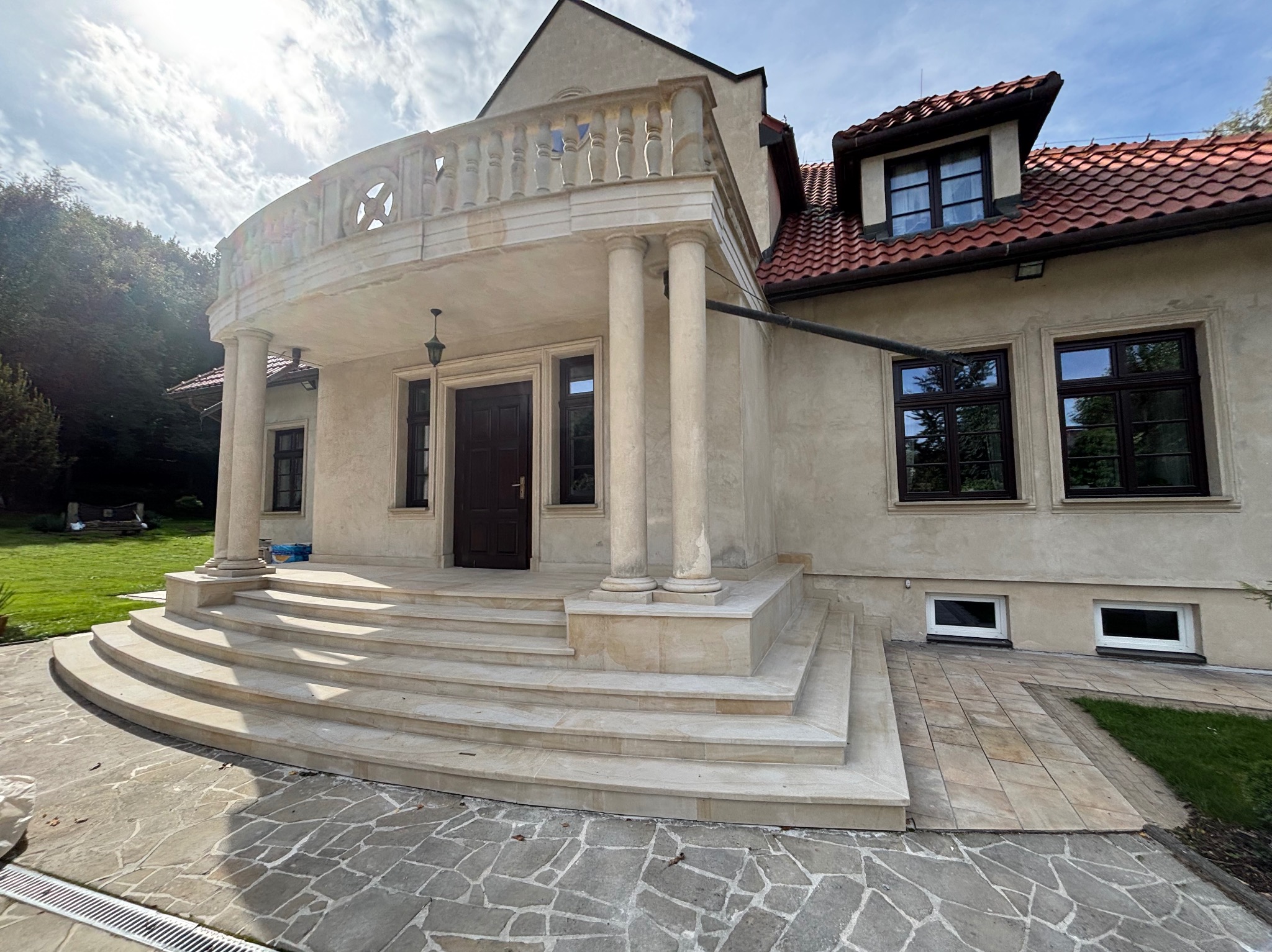 Eleganckie schody kamienne prowadzą do wejścia domu z kolumnami i balkonem. Kamienna kostka brukowa na podjeździe. Całość w jasnych, stonowanych barwach.