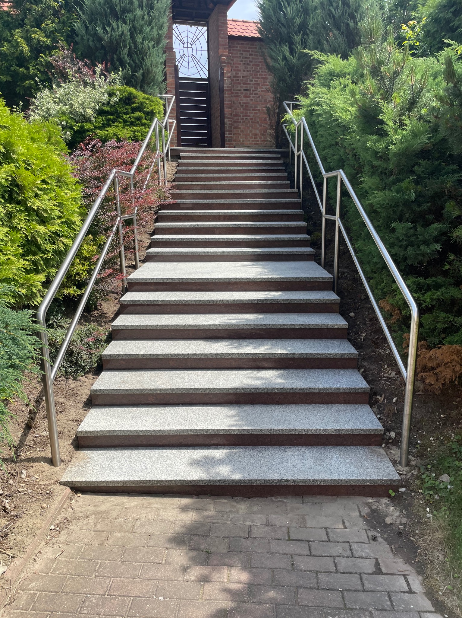 Zewnętrzne schody z szarego kamienia z metalową balustradą, prowadzące do budynku przez bujny ogród z różnorodną roślinnością. Ujęcie z dołu.