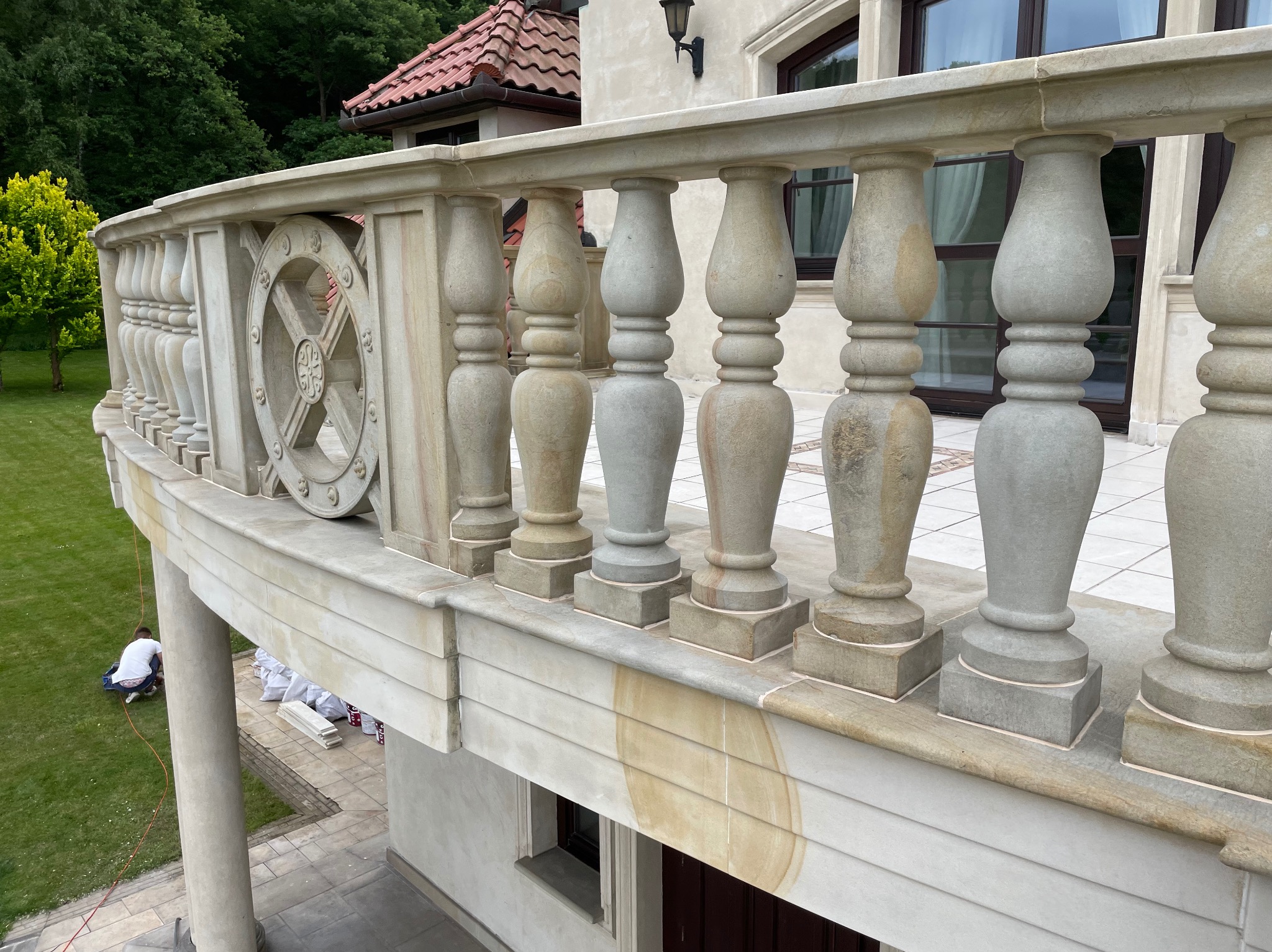 Kamienna balustrada balkonu z dekoracyjnymi elementami, widok z góry na trawnik i pracownika w trakcie montażu. Widoczne detale wykończenia i faktura kamienia.