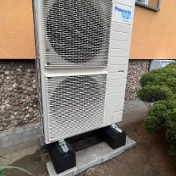 Zewnętrzna jednostka pompy ciepła Panasonic, zamontowana na betonowej podstawie z czarnymi podkładkami antywibracyjnymi, na tle ściany budynku z kamienną okładziną i oknem.