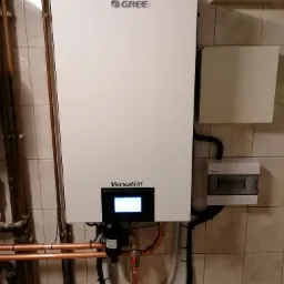 Biały, wiszący na ścianie piec gazowy Gree Versati III z podłączonymi miedzianymi rurami i czarnym filtrem, na tle beżowych płytek ceramicznych.