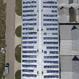 Widok z g&oacute;ry na instalację fotowoltaiczną na dachu hali przemysłowej, obok droga i zaparkowany bus serwisowy, oraz sąsiednie budynki z charakterystycznymi świetlikami dachowymi.