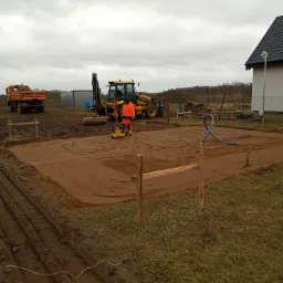 Pracownik w pomarańczowej odzieży roboczej ubija ziemię zagęszczarką wibracyjną na placu budowy, w tle koparko-ładowarka Volvo, wywrotka ZURG, oraz fundamenty domu jednorodzinnego.