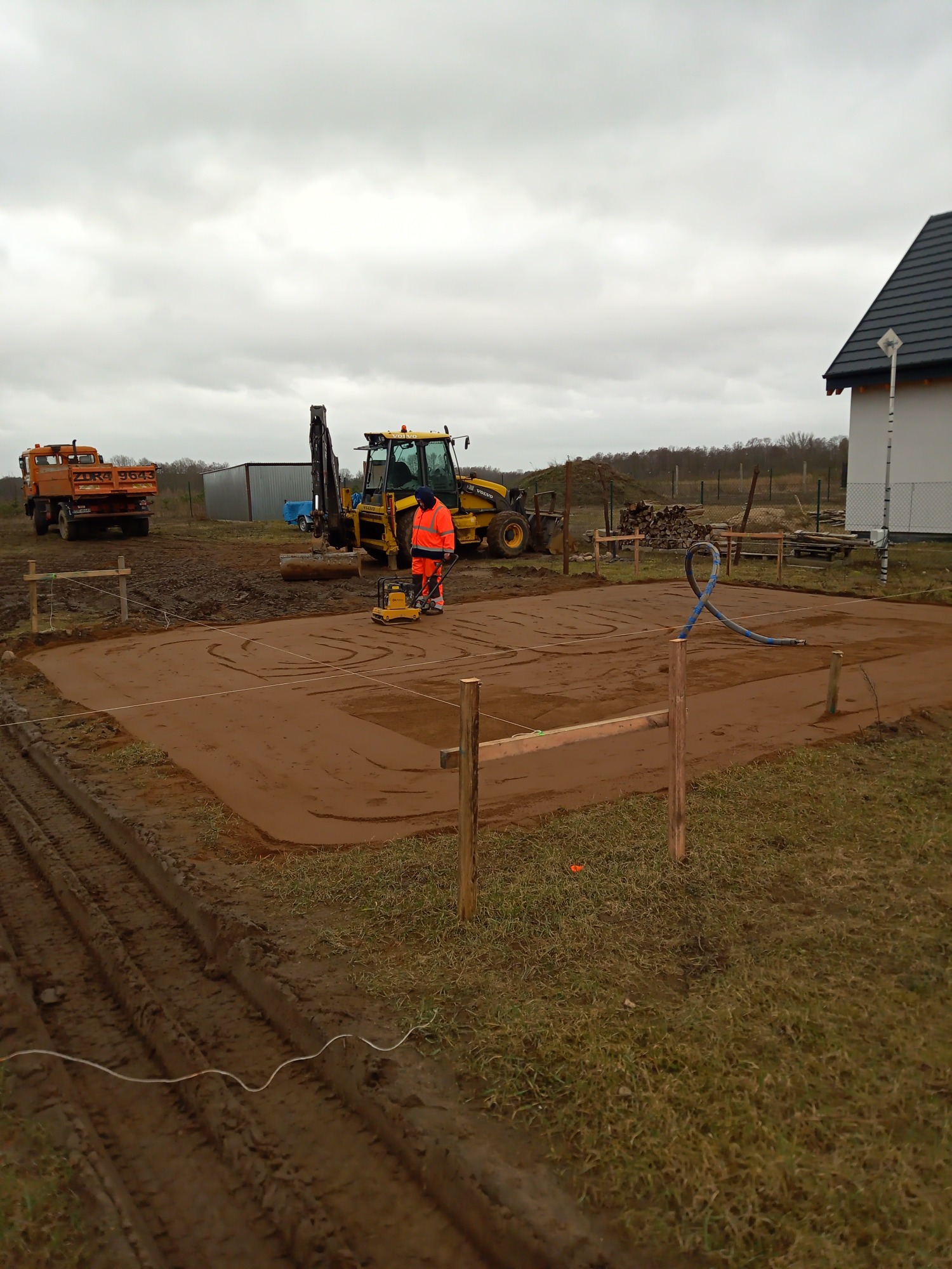 Pracownik w pomarańczowej odzieży roboczej ubija ziemię zagęszczarką wibracyjną na placu budowy, w tle koparko-ładowarka Volvo, wywrotka ZURG, oraz fundamenty domu jednorodzinnego.