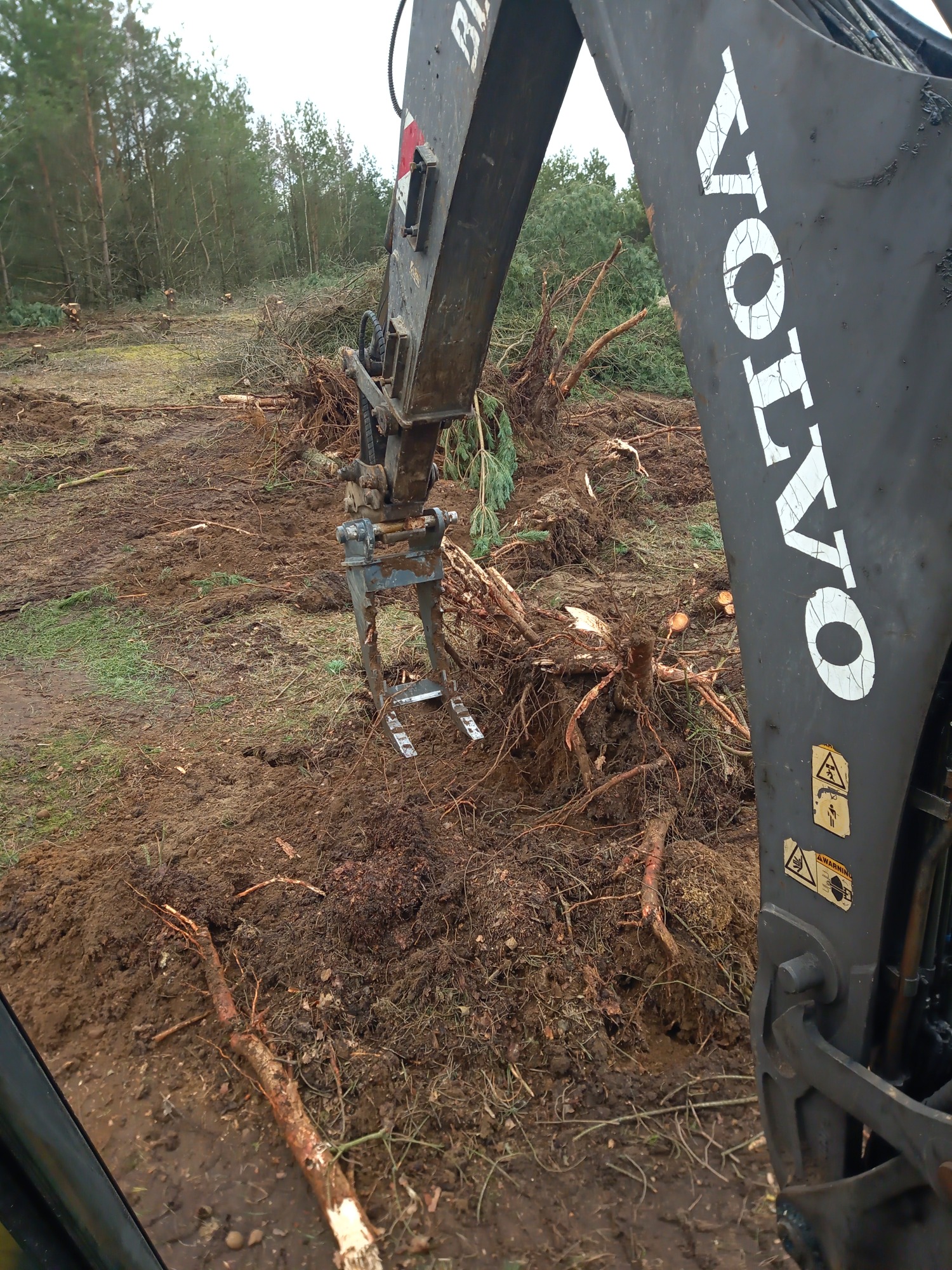 Ciężki sprzęt marki Volvo, z chwytakiem do pni, usuwa korzenie i gałęzie z terenu przygotowywanego pod inwestycję, widoczne ścięte pnie drzew na horyzoncie.