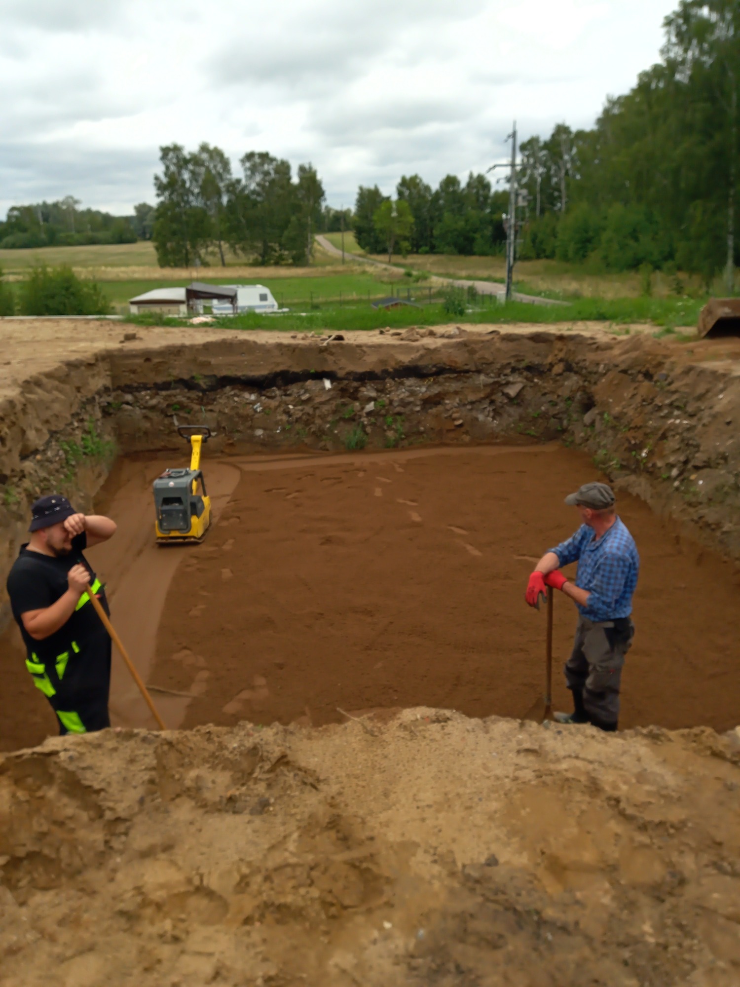 Dwaj pracownicy w wykopie wyrównują teren ubijarką wibracyjną, przygotowując podłoże pod fundamenty, w tle widoczny krajobraz wiejski.