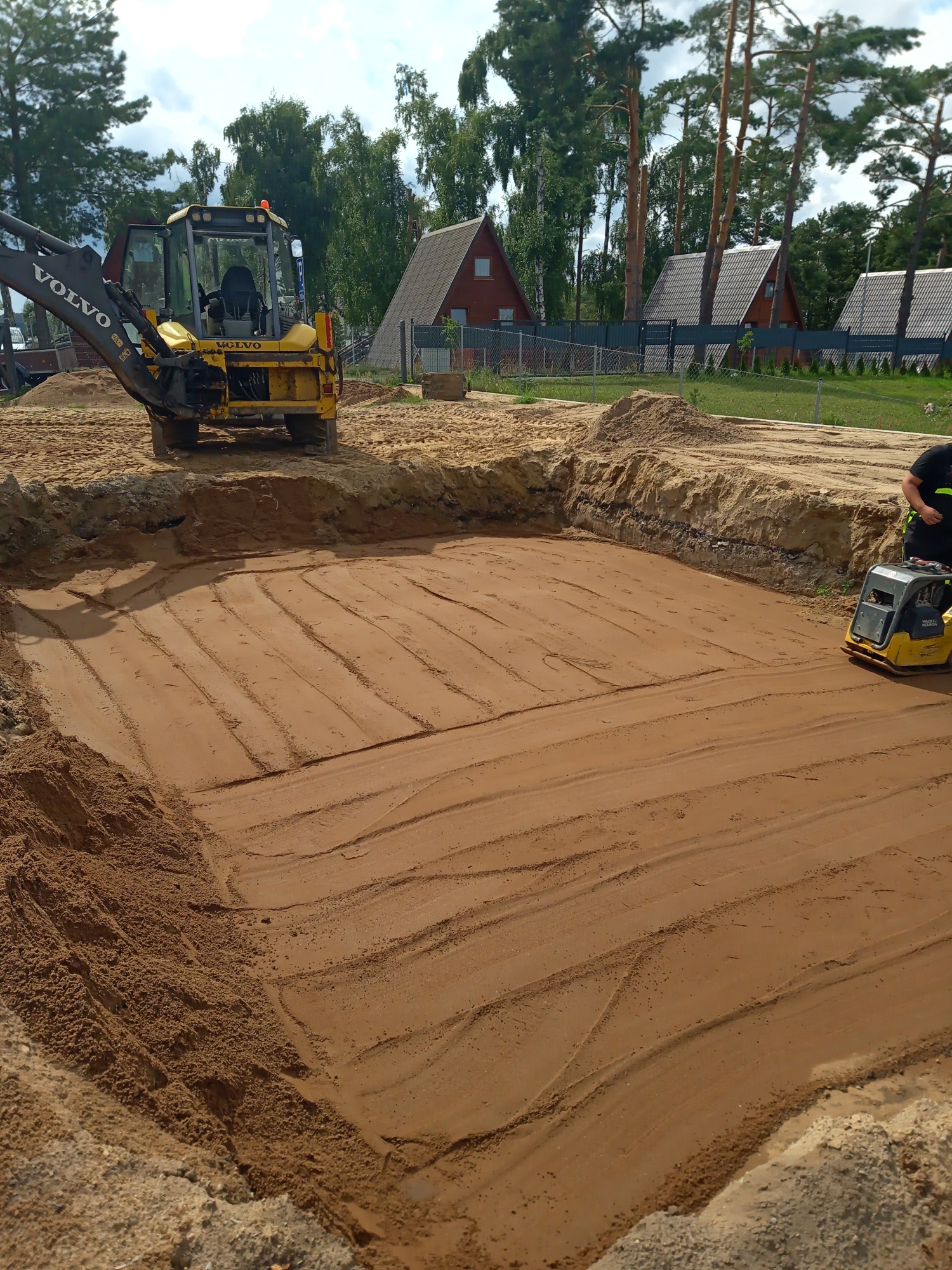Żółta koparko-ładowarka Volvo i zagęszczarka w wykopie wypełnionym piaskiem, przygotowywanym pod fundamenty budynku, w tle domy jednorodzinne i drzewa.