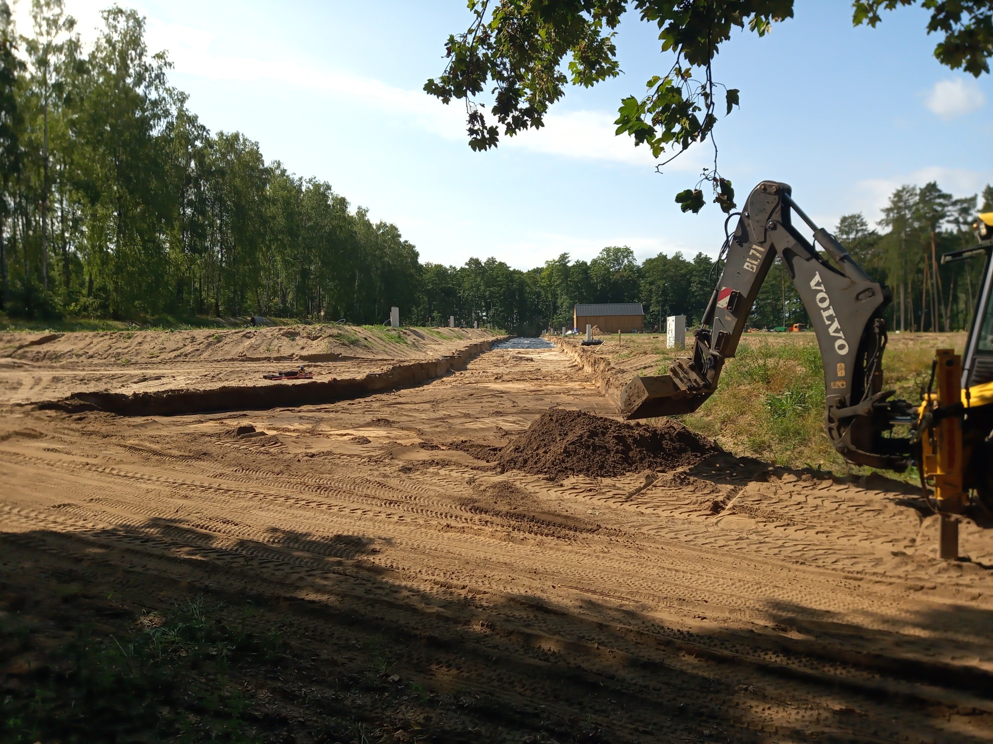 Żółta koparka Volvo BL71 przy wykopie w piaszczystym terenie, z widocznymi śladami opon i stertą ziemi, w tle drzewa i budynek.