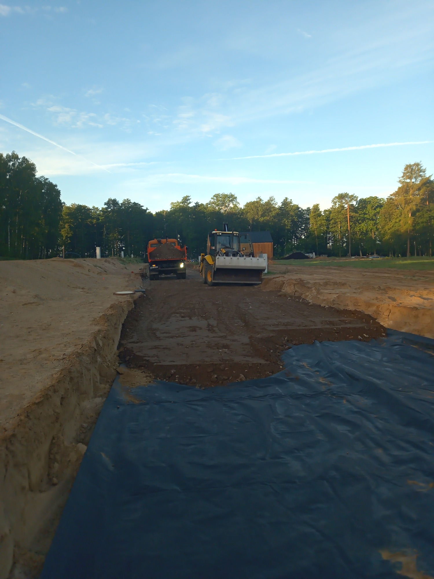 Żółta równiarka i pomarańczowa wywrotka pracują na placu budowy z wykopem zabezpieczonym czarną folią, w tle drzewa i błękitne niebo.