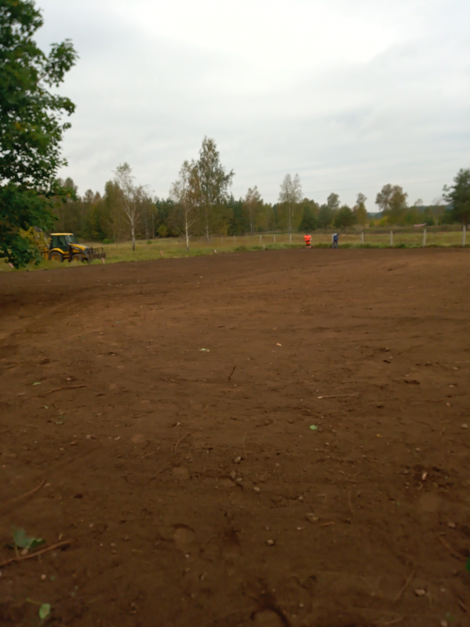 Przygotowany, brązowy teren w Czaplinku, z widocznym traktorem z bronami w tle oraz pracownikami przy ogrodzeniu.