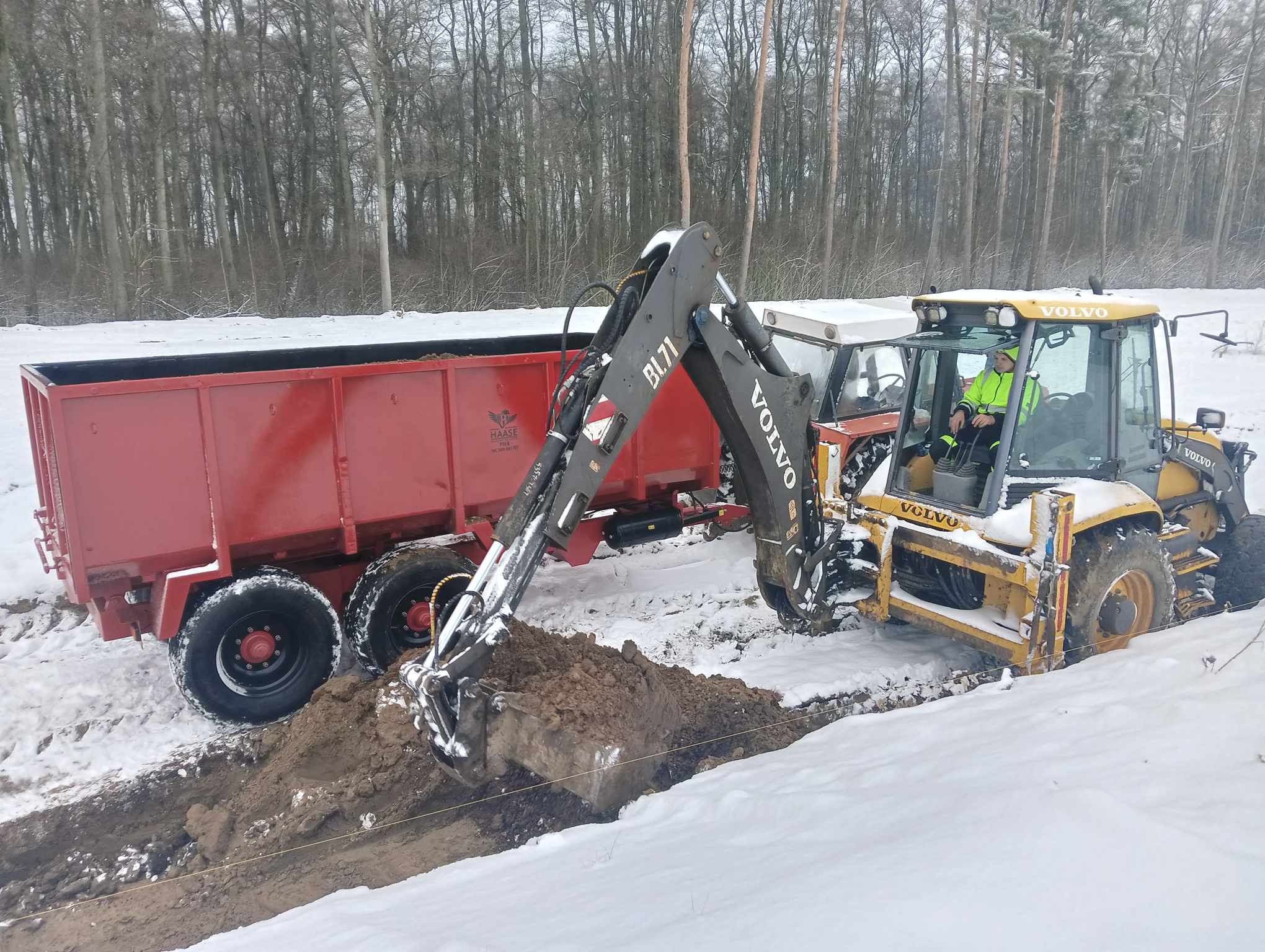 Żółta koparko-ładowarka Volvo BL71 kopie rów w zasypanym śniegiem terenie, obok stoi czerwona przyczepa. W tle widoczny las.