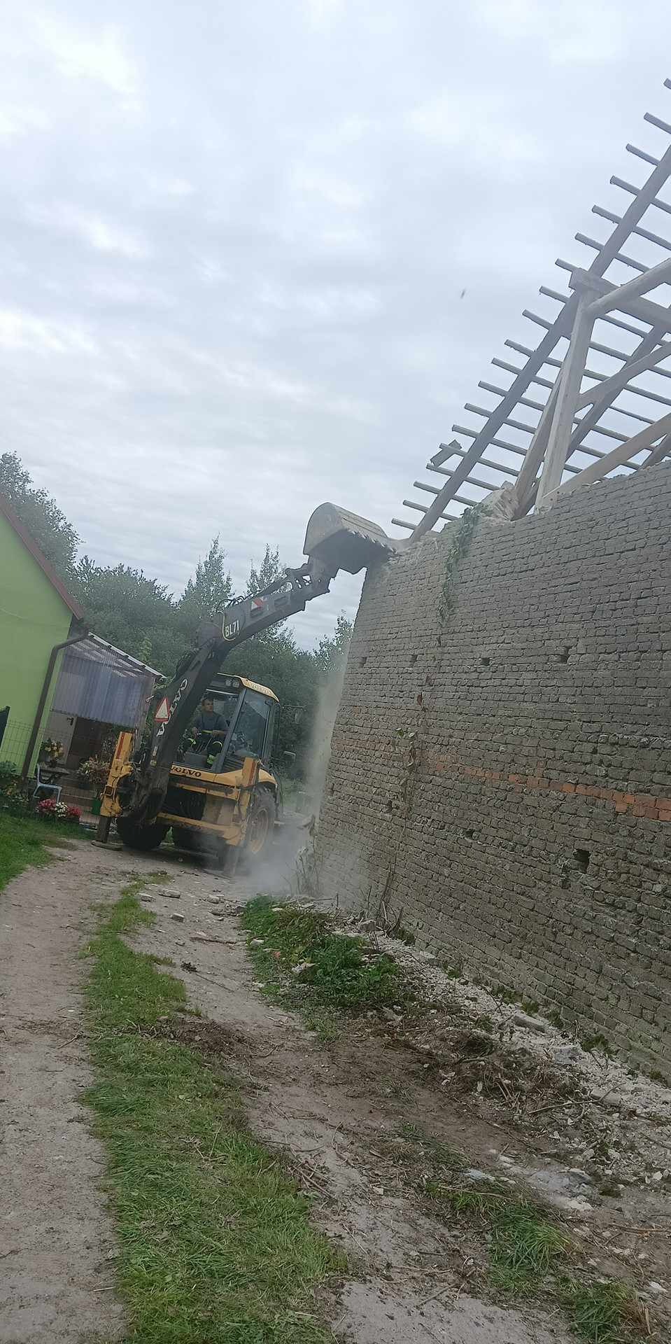 Żółta koparko-ładowarka Volvo BL71 kruszy ceglany mur starego budynku z drewnianą konstrukcją dachu, pył i gruz w powietrzu, zielony budynek w tle.