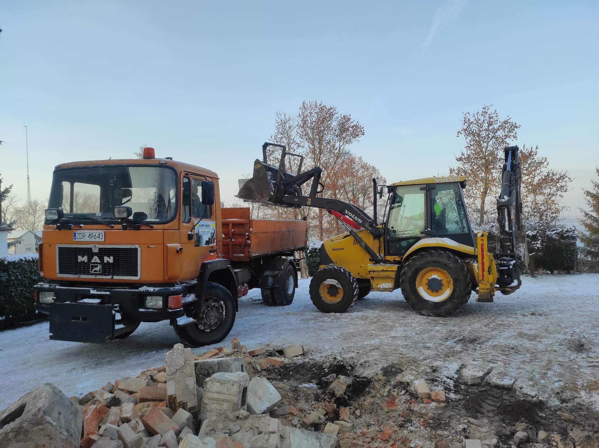 Pomarańczowy samochód ciężarowy MAN z wywrotką oraz żółta koparko-ładowarka Volvo na placu z gruzem ceglanym i resztkami śniegu.