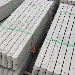 słupki ogrodzeniowe betonowe ,,Mar-Bud'' - Stosy prefabrykowanych słupków betonowych do ogrodzeń, zabezpieczone zieloną taśmą, ułożone na paletach, gotowe do transportu.