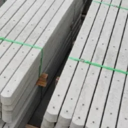 Szare betonowe słupki do siatki ogrodzeniowej ułożone w stosy, spięte zielonymi taśmami, widoczne otwory montażowe.