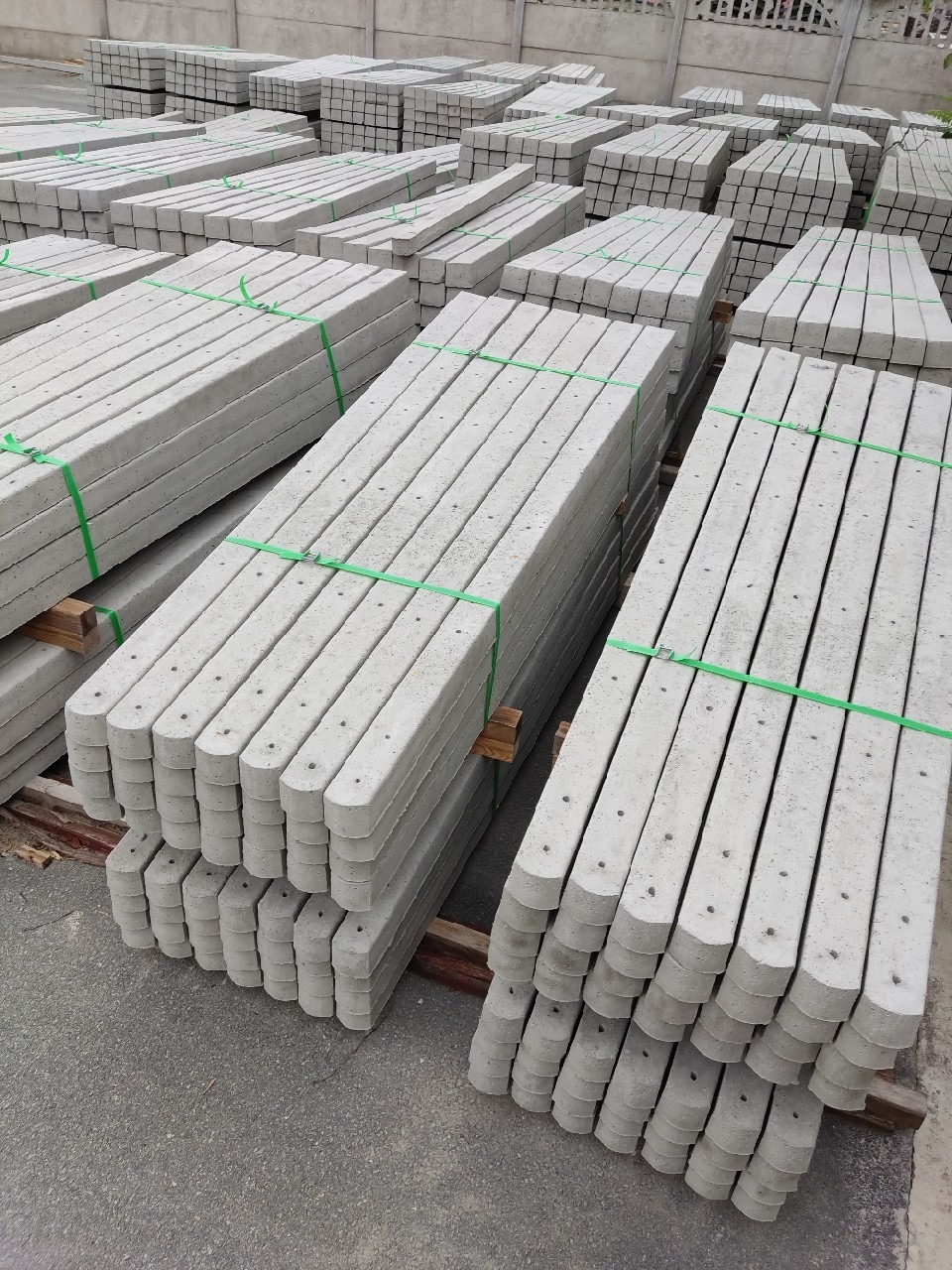 Stosy prefabrykowanych słupków betonowych z otworami, zabezpieczone zielonymi taśmami na drewnianych paletach, przygotowane do transportu lub montażu.